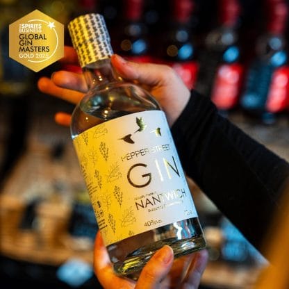 A person holds a bottle of Pepper Street Gin with a white and gold label featuring botanical illustrations and bird silhouettes. A “Global Gin Masters Gold 2025” award badge is shown in the top left corner. Bottles on a shelf are blurred in the background.