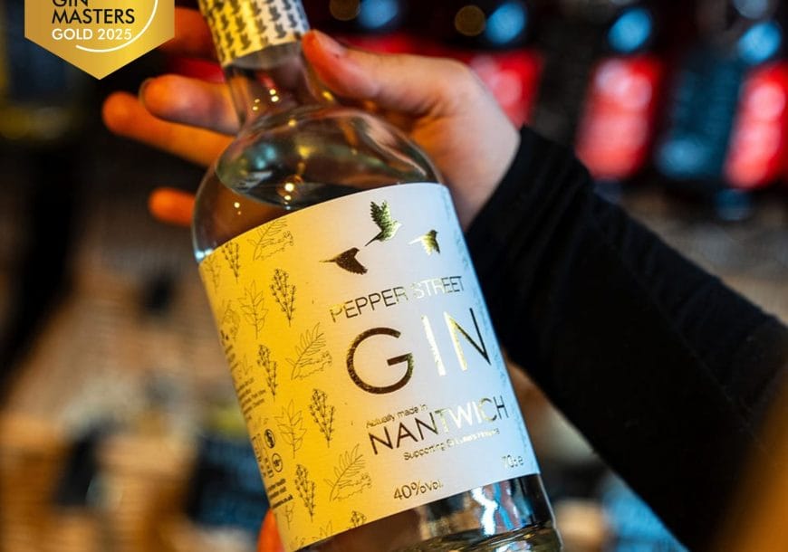 A person holds a bottle of Pepper Street Gin with a white and gold label featuring botanical illustrations and bird silhouettes. A “Global Gin Masters Gold 2025” award badge is shown in the top left corner. Bottles on a shelf are blurred in the background.