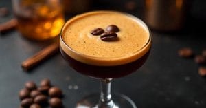 A dark espresso martini in a coupe glass, topped with a creamy foam and garnished with three coffee beans, surrounded by scattered coffee beans and a cinnamon stick.