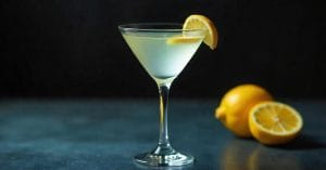 A martini glass filled with a pale yellow cocktail, garnished with a lemon slice on the rim, set on a dark surface with two lemons, one halved, in the background.