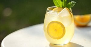 A clear glass of lemonade with ice cubes, lemon slices, and a sprig of mint, set on a white table with a blurred green background.