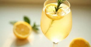 A light golden cocktail served in a wine glass, garnished with a lemon slice and a sprig of rosemary, with halved lemons and soft natural lighting in the background.