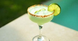 A pale green frozen margarita served in a stemmed cocktail glass with a spiced rim, garnished with a lime wheel and a twist of lime peel, placed beside a pool.