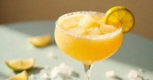 A yellow frozen cocktail served in a coupe glass with a salted rim, ice cubes, and a lemon wheel garnish. Lemon wedges and ice cubes are scattered on the table in the background.