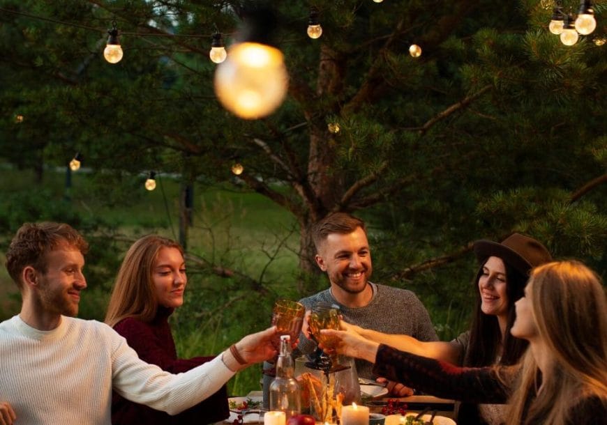A group of six friends sitting around an outdoor dinner table at dusk, toasting with drinks under string lights and surrounded by trees.