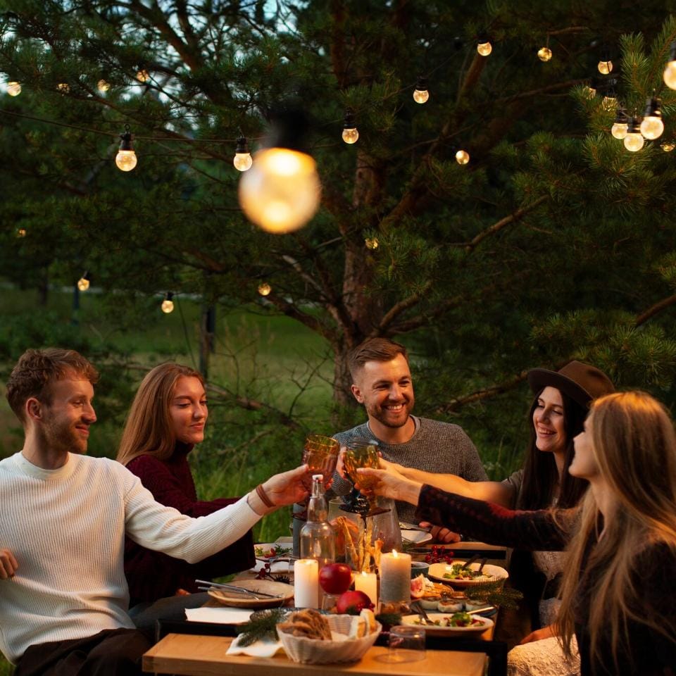 A group of six friends sitting around an outdoor dinner table at dusk, toasting with drinks under string lights and surrounded by trees.