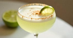 A frothy pale green lime cocktail in a coupe glass with a graham-cracker rim, garnished with a lime wheel on the rim and sprinkled crumbs on the foam, with a blurred halved lime in the background.