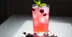 A tall glass filled with a pink iced beverage, garnished with floating cranberries and a mint sprig, placed on a folded napkin on a softly lit table.