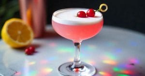 A pink cocktail in a coupe glass with a foamy top, garnished with red berries and a lemon twist, sitting on a surface with rainbow light reflections.