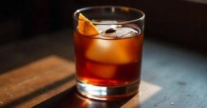 A glass of dark iced coffee with a large ice cube, garnished with an orange peel and a coffee bean, on a wooden and dark surface in moody lighting.