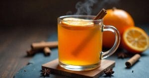 A steaming glass mug filled with hot orange-hued beverage, garnished with a cinnamon stick and an orange slice, surrounded by whole oranges, cinnamon sticks, and star anise.