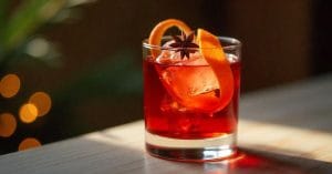 A short glass with a red cocktail, garnished with ice cubes, a twist of orange peel, and a star anise on top.
