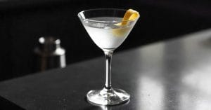 A clear martini glass filled with a transparent cocktail, garnished with a twist of lemon peel, placed on a sleek dark surface.