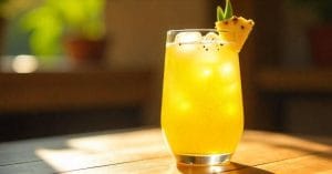 A tall glass of yellow tropical drink with ice, garnished with a pineapple wedge and leaf, sitting on a sunlit wooden surface.
