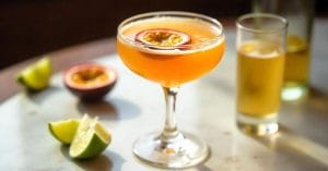 A cocktail glass filled with a vibrant orange drink garnished with half a passion fruit. Surrounding the glass are lime wedges, a halved passion fruit, and a tall glass with a golden beverage in the background, all set on a sunlit white marble table.