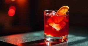A dark red cocktail served in a short glass with ice, garnished with an orange slice, set against a dimly lit background with warm, blurry red lights.
