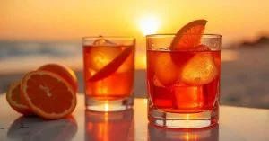 Two glasses of orange cocktails with ice and orange slices, set on a table by the beach at sunset, with fresh orange halves beside them and a warm golden glow in the background.