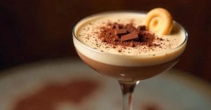 A creamy layered dessert drink in a cocktail glass, topped with cocoa powder, chocolate chunks, and a rolled cookie, against a warm blurred background.