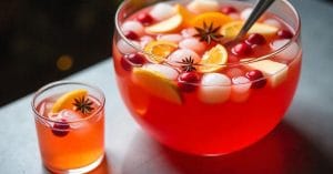 A large glass punch bowl filled with red fruit punch, ice, orange slices, cranberries, star anise, and a ladle, with a matching glass of punch beside it.